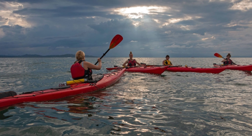 A group of people paddle individual red kayaks on open water under gray skies. 